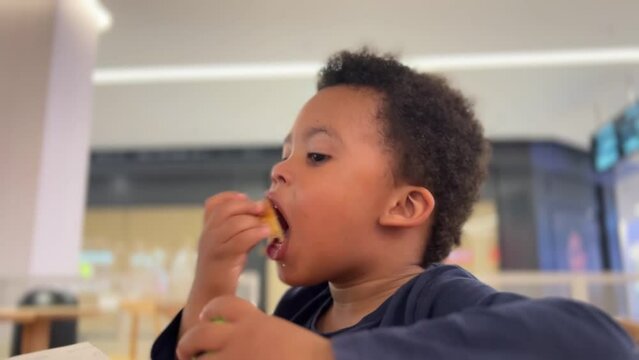 Exotic black 3-year-old child enjoying eating a cake in a cafeteria. He wears a long sleeve blue shirt.
