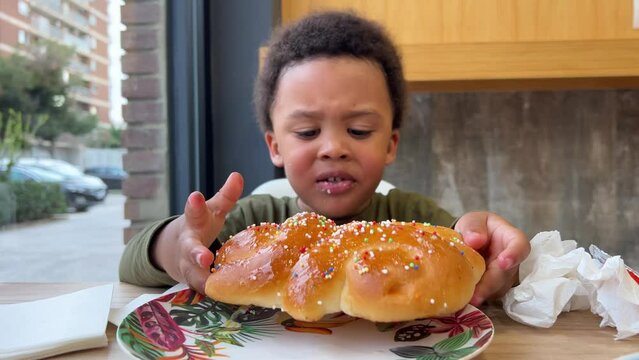 Three-year-old happy black child eating a big cake in a cafeteria seated next to his mother. The cake is a traditional spanish caka &ldquo;mona de Pascua&rdquo;.