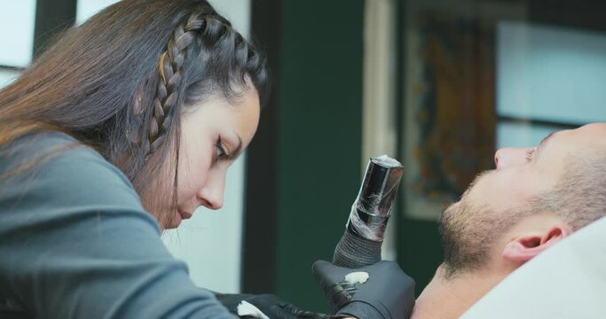A young female tattoo artist is tattooing the chest of a young man in her tattoo studio, using the dotwork technique