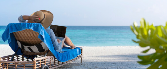 A young woman works on a laptop on the beach in the Maldives. The concept of remote work and nomadism. Banner