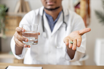 Focus on green round pill being held by multiracial male doctor sitting at writing desk with laptop on office background. Professional medical worker giving medicine and glass of water for treatment.