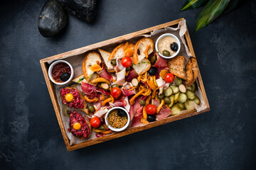 Banquet meat plate with slices of ham, olives, cherry tomatoes, honey mushrooms, pickled cucumbers, tar tar, mustard and bread on a dark background.