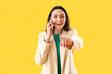 Happy young woman talking by phone and pointing at viewer on yellow background