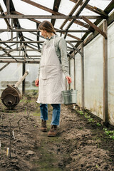 An attractive woman holding a tin bucket and walking in the greenhouse looking at seedlings