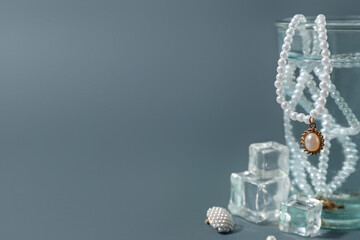 Glass of water with stylish jewelry and ice cubes on grey background