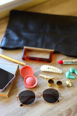 Handbag, sunglasses, beauty products, pearl hair clips, rings, pen, earbuds, book and phone on wooden background. Selective focus.