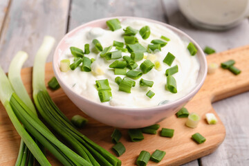 Bowl of tasty sour cream with green onion on light wooden background, closeup