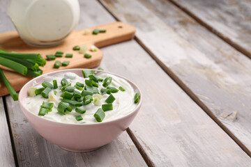 Bowl of tasty sour cream with green onion on light wooden background