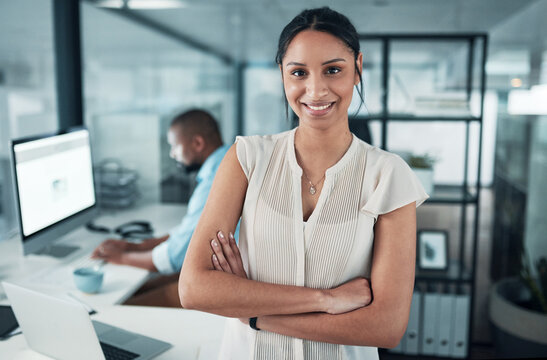 Being young in a corporate world takes strength. an attractive young businesswoman standing with her arms folded while a colleague works behind her.