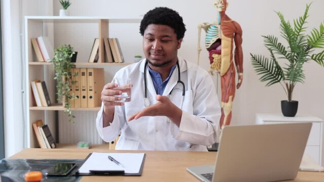 Positive multicultural physician in lab coat showing glass of water while sitting at writing desk with laptop and documents in doctor's office. General practitioner recommending staying hydrated.