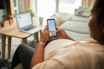 Young pregnant latina woman using a diet app on her smart phone while sitting on a couch in the living room of her home