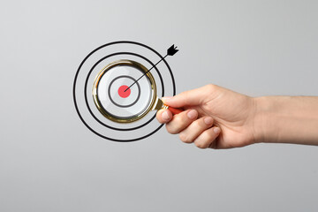 Woman holding magnifying glass on light grey background, closeup. Illustration of target and arrow