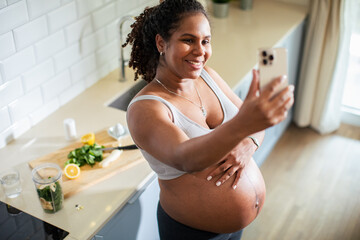 Young pregnant latina woman using a smart phone and having a video call while in the kitchen of a...