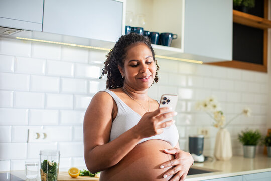 Young pregnant latina woman using a smart phone in the kitchen of a home - Powered by Adobe