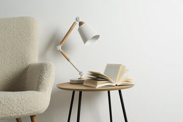 Stylish lamp with open book on wooden coffee table and soft armchair near white wall. Interior...