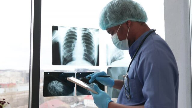 Attentive multiethnic man in medical scrubs and face mask looking at X-ray pictures while holding tablet in doctor's workplace. Physician in gloves and cap examining chest scans in hospital interior.
