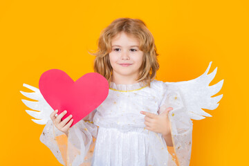 Child angel pointing on paper red heart, point gesture. Child angel. Portrait of cute kid with angel wings isolated on yellow studio background. Little angel, valentines day. Angelic kids.