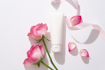 Advertising scene for cosmetics with rose extract. White plastic tube container cream, gel for washing with ribbon, roses and rose petals on white background. Natural pure natural cosmetics