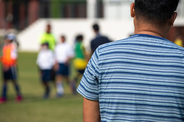 Father standing and watching his son playing football in a school tournament on a clear sky and...