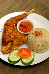 Ayam Goreng is indonesian fried chicken, served with brown rice, fresh vegetables such as tomato and cucumber, and chili sauce. On a brown wooden table. Indonesian food. 