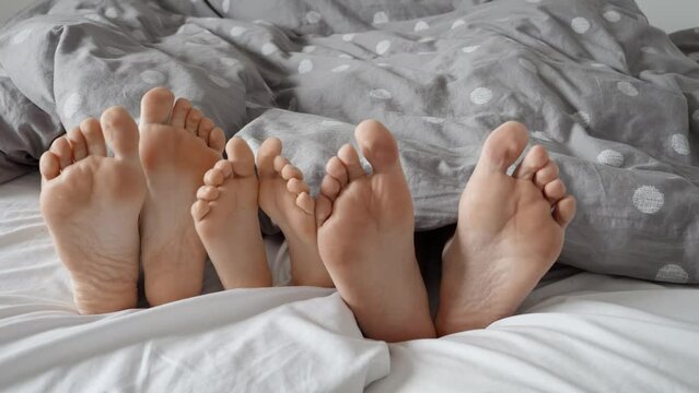 Closeup of happy man's, women's and child's feet lying on soft bed and showing under the blanket. Concept of family love, resting and relaxing at home together, relationship and family life, parenting