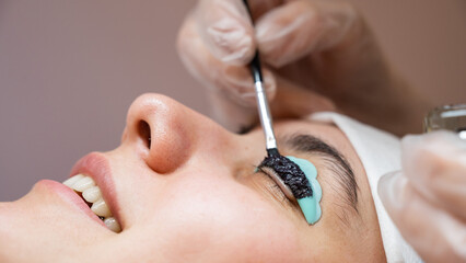 Close-up portrait of a woman on eyelash lamination procedure. The master applies tint to the...