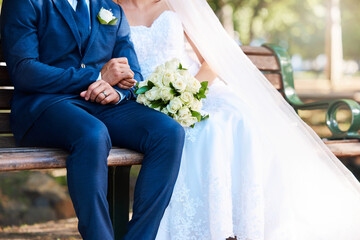 Closeup of a bride and groom sitting on a park bench together. Man wearing suit and woman wearing...