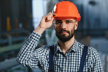 Portrait of Professional Heavy Industry Engineer Worker Wearing Safety Uniform, Hard Hat Smiling. In the Background Unfocused Large Industrial Factory.