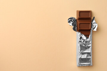 Delicious chocolate bar wrapped in foil on beige background, top view. Space for text