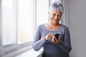 Window, app and senior woman with a smartphone, connection and typing for social media at home. Mature lady, female person and model with a cellphone, communication and online reading with texting