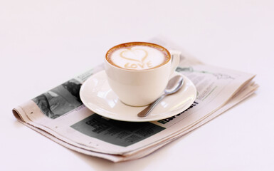 Coffee cup, cappuccino and newspaper with heart in foam on a table with no people in a restaurant. Cafe drink, love writing and foamy beverage art with milk in a latte and a closeup of mug and spoon