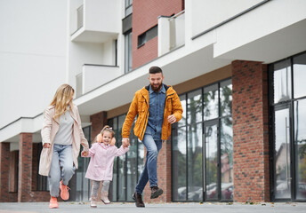 Full length of man and woman holding hands of little girl while walking down the street in new urban district. Happy loving family with child enjoying stroll outdoors near residential building. - obrazy, fototapety, plakaty