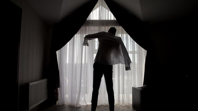 A handsome man in his loft dresses his sexy white shirt in a sexy way, looks into his room and smiles with a sure look at himself. Concept of: elegance, business, work.
