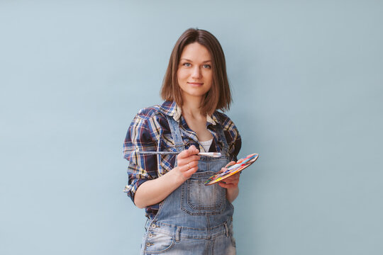 The artist in a plaid shirt on a blue background holds a paint palette and a brush. Creative person concept. She is wearing denim overalls and a plaid shirt.