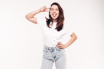 Fototapeta premium Young beautiful woman. Trendy carefree model in casual summer white T-shirt and jeans. Positive female isolated on white in studio. Cheerful and happy. Copy space, mockup. Shows tongue, peace sign
