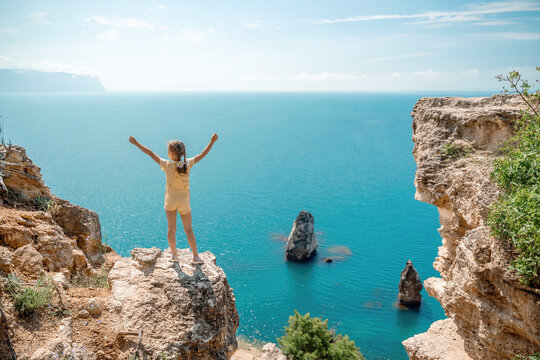 Happy girl stands on a rock high above the sea, wearing a yellow jumpsuit and sporting braided hair, depicting the idea of a summer vacation by the sea.