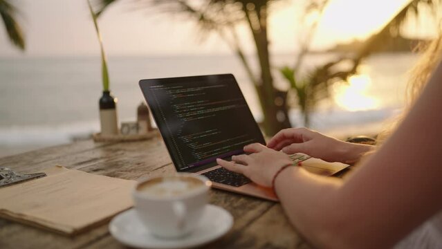 Young female developer working on laptop by the ocean. Woman freelancer coding at outdoor tropical cafe at sunset. Caucasian girl working remotely typing on computer at exotic location. Worldwide work
