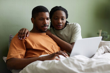Fototapeta premium African American young couple watching movie on laptop while resting in bed in bedroom