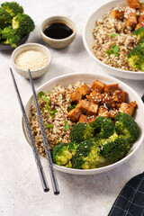 One quinoa bowl with steamed broccoli and smoked tofu cubes in teriyaki sauce with sesame seeds as topping, black chopsticks, grey concrete background