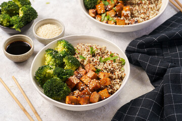 Two quinoa bowls with steamed broccoli and smoked tofu cubes in teriyaki sauce with sesame seeds as topping on white checkered kitchen napkin, grey concrete background