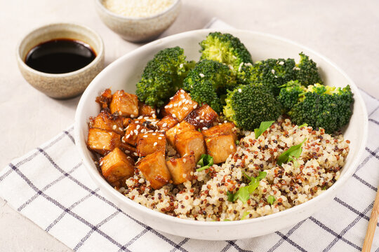 One quinoa bowl with steamed broccoli and smoked tofu cubes in teriyaki sauce with sesame seeds as topping on white checkered kitchen napkin, grey concrete background