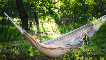 Young happy caucasian woman in a hat lying in a hammock in a green garden enjoying a summer holidays