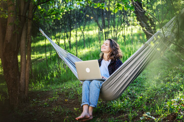 Young cheerful dreamy woman sitting with laptop in hammock in summer garden, countryside lifestyle...