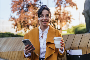 Fototapeta premium Portrait of a business woman on a coffee break listening to a podcast in a park,drinking coffee. Woman sitting on a bench in a park looking away from camera, it's fall and she's wearing a coat