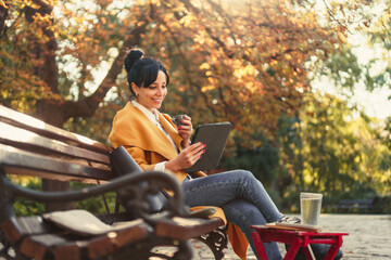 Happy young girl sitting in the park drinking coffee and reading. Beautiful woman having video call.. Woman in a park with tress in the background