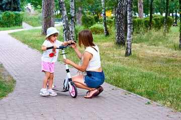 A young mother raises educates a child with a scooter on a path in a summer park