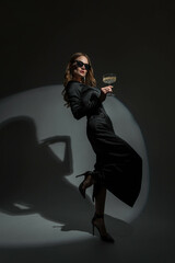 Fototapeta Gorgeous beautiful stylish woman with cool glasses in a trendy black evening dress with heels holds a glass of champagne and celebrates the event on a dark background. Pretty chic Lady at the party obraz