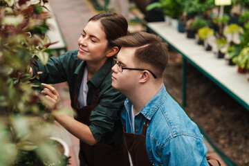 Woman florist helping young man with down syndrome to handle with flowers in greenhouse