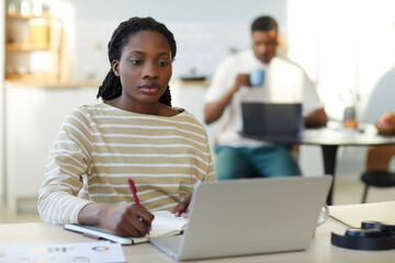 African American woman making notes in notebook while working online on laptop sitting at table at home