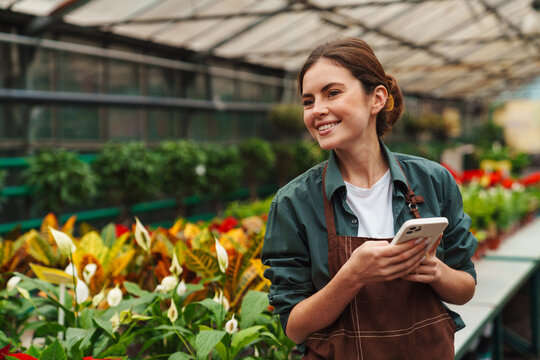 Smiling woman gardener using mobile phone while working in greenhouse
